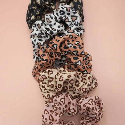 Set of five leopard pattern scrunchies in neutral tones arranged on a blush pink background, highlighting their soft, stretchy polyester fabric and playful prints.