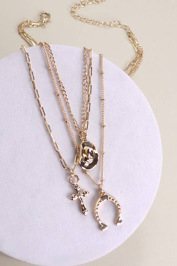 Gold plated multi-layer western inspired charm necklace with dangling horseshoe, porcelain hat and cross charms styled on pink background.