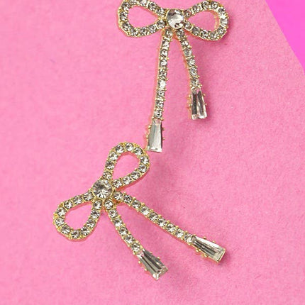 Gold rhinestone bow drop earrings with a delicate dangling design, shown worn on the ear and displayed against a bright pink background.