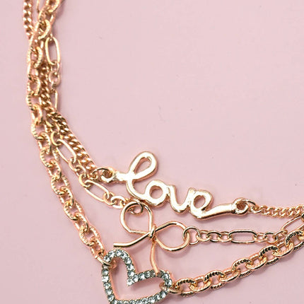 Close-up of a gold-plated layered charm bracelet featuring a rhinestone heart, bow charm, and “LOVE” letter charm displayed on a soft pink background.