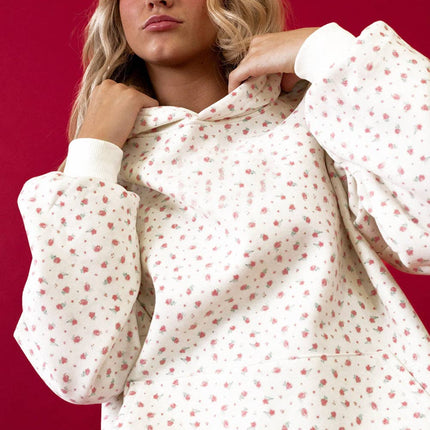 Model wearing a pink floral hoodie with a kangaroo pocket and matching sweatpants styled in a relaxed fit against a red background.
