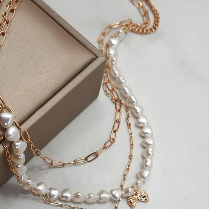 Gold layered necklace featuring pearl strand, bow charm, and rhinestone heart pendant styled on a neutral background