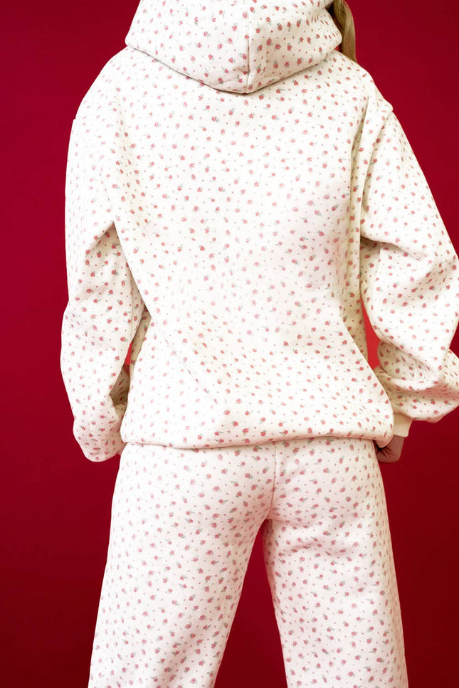 Model wearing a pink floral hoodie with a kangaroo pocket and matching sweatpants styled in a relaxed fit against a red background.