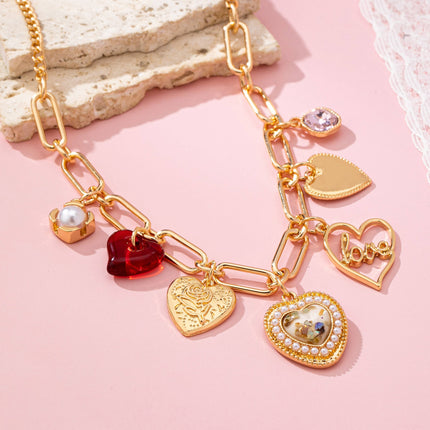Gold plated elongated paperclip chain necklace with dangling porcelain heart, rhinestone crystal, pearl cluster, and red enamel heart charms styled in a romantic feminine jewelry aesthetic.