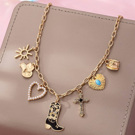 Gold paperclip chain necklace with western-inspired charms including a cowboy boot, heart, cross, sunburst, and hat, styled on a model’s neckline against a soft neutral background