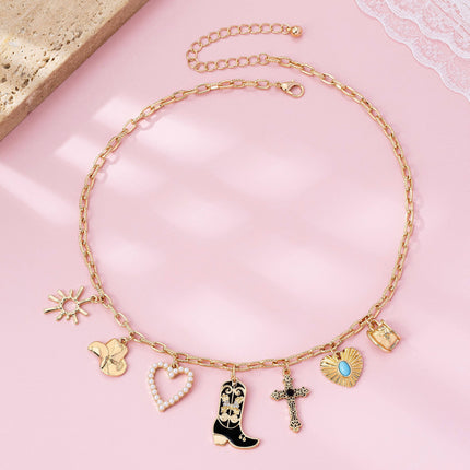 Gold paperclip chain necklace with western-inspired charms including a cowboy boot, heart, cross, sunburst, and hat, styled on a model’s neckline against a soft neutral background