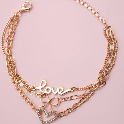 Close-up of a gold-plated layered charm bracelet featuring a rhinestone heart, bow charm, and “LOVE” letter charm displayed on a soft pink background.