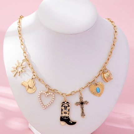 Gold paperclip chain necklace with western-inspired charms including a cowboy boot, heart, cross, sunburst, and hat, styled on a model’s neckline against a soft neutral background