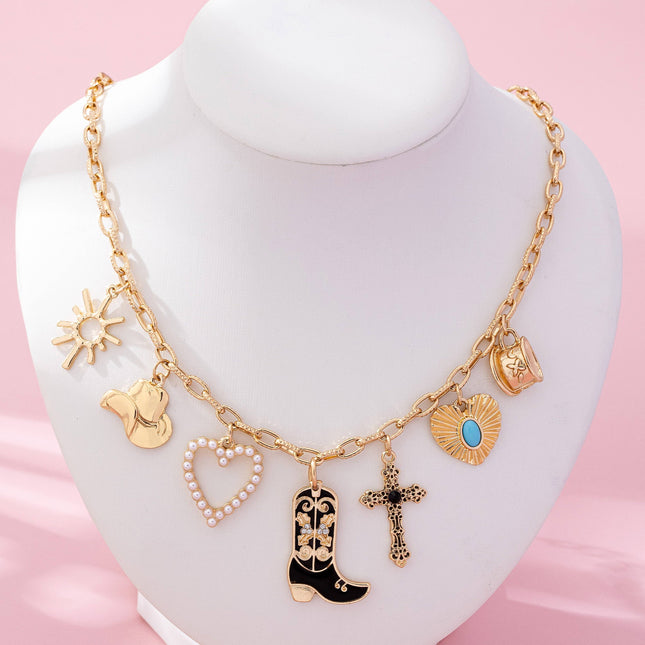 Gold paperclip chain necklace with western-inspired charms including a cowboy boot, heart, cross, sunburst, and hat, styled on a model’s neckline against a soft neutral background