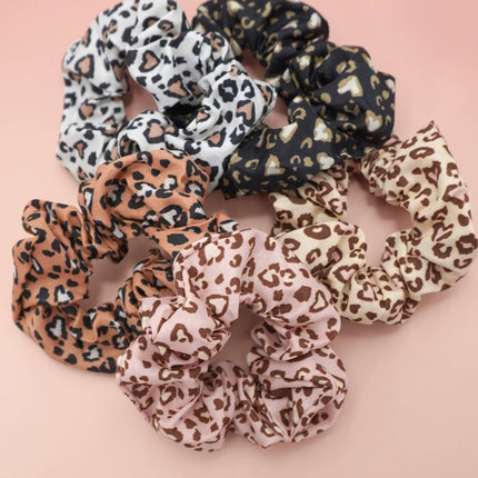 Set of five leopard pattern scrunchies in neutral tones arranged on a blush pink background, highlighting their soft, stretchy polyester fabric and playful prints.