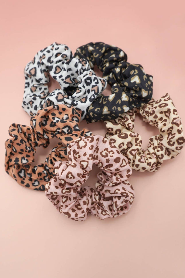 Set of five leopard pattern scrunchies in neutral tones arranged on a blush pink background, highlighting their soft, stretchy polyester fabric and playful prints.
