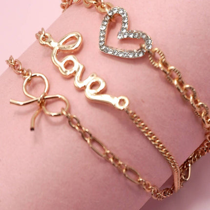 Close-up of a gold-plated layered charm bracelet featuring a rhinestone heart, bow charm, and “LOVE” letter charm displayed on a soft pink background.