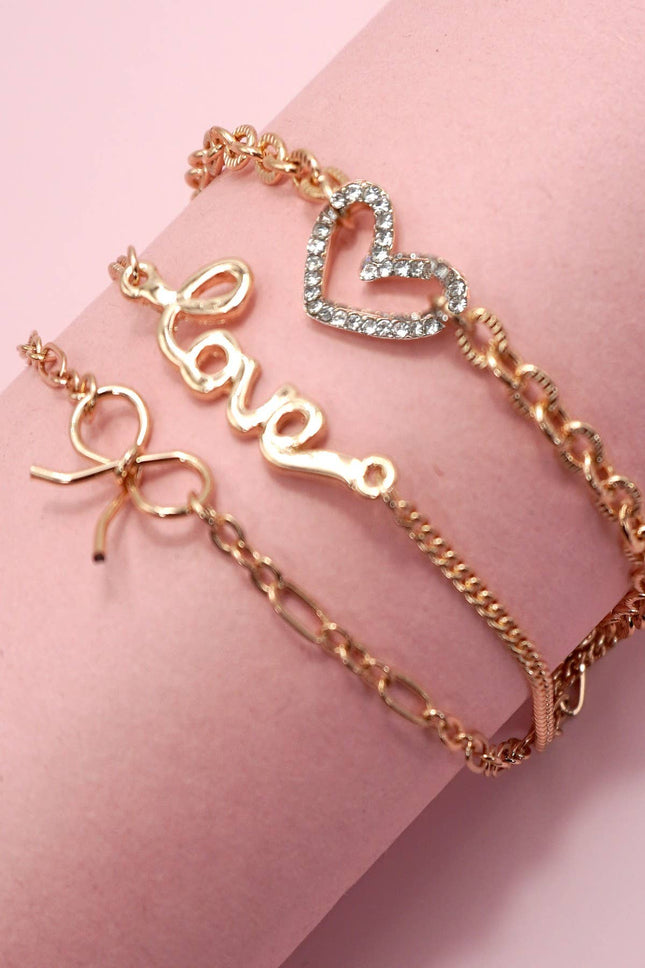 Close-up of a gold-plated layered charm bracelet featuring a rhinestone heart, bow charm, and “LOVE” letter charm displayed on a soft pink background.