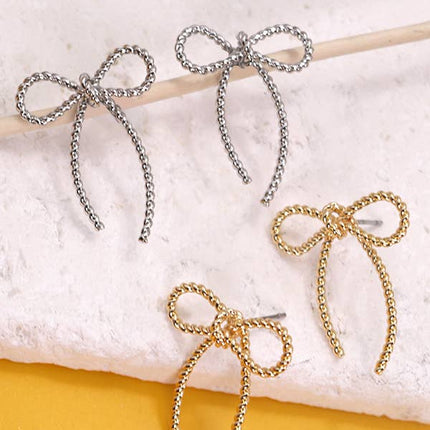 Bow-shaped earrings in gold and silver on a textured surface with a yellow background
