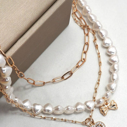 Gold layered necklace featuring pearl strand, bow charm, and rhinestone heart pendant styled on a neutral background