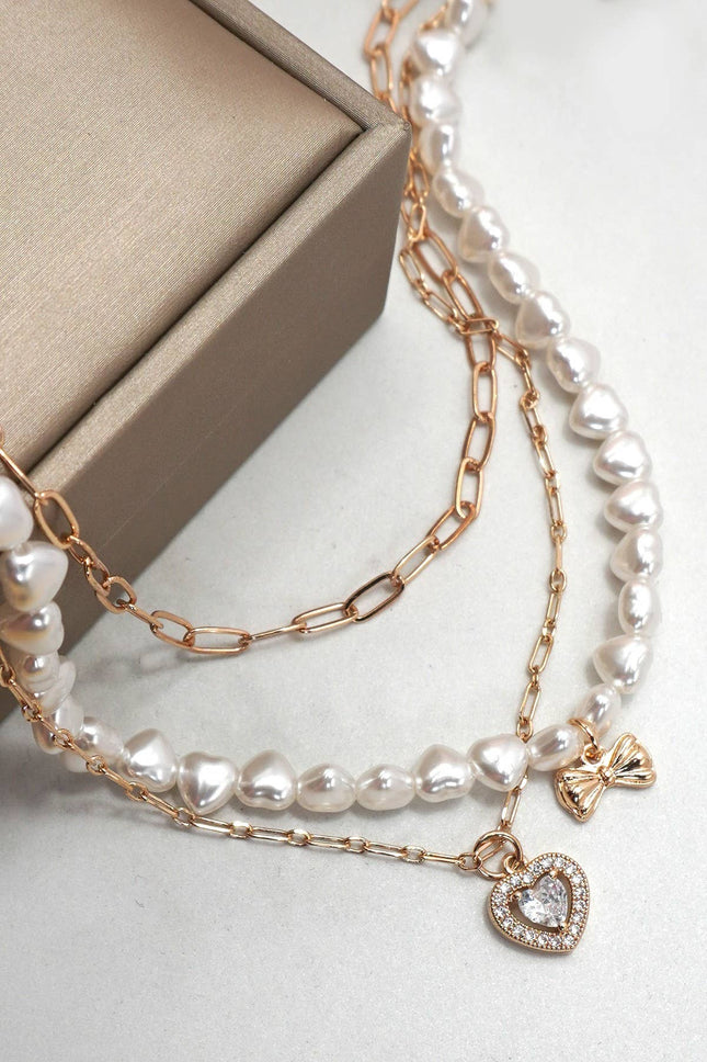 Gold layered necklace featuring pearl strand, bow charm, and rhinestone heart pendant styled on a neutral background