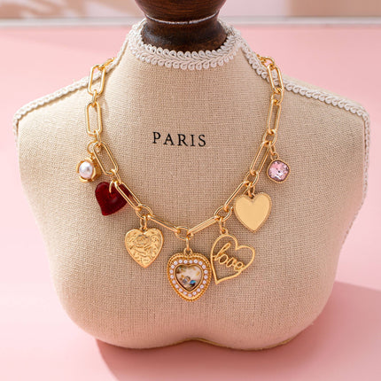 Gold plated elongated paperclip chain necklace with dangling porcelain heart, rhinestone crystal, pearl cluster, and red enamel heart charms styled in a romantic feminine jewelry aesthetic.