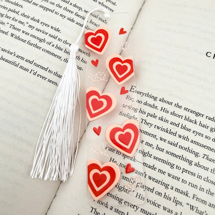 Handmade acrylic Valentine’s Day bookmarks featuring pink and red heart-themed designs with tassels, styled on open books for a cozy, festive reading aesthetic.