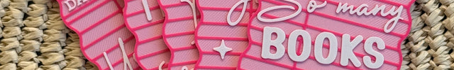 Five pink paper library card bookmarks with white printed text and lines, arranged overlapping on a woven straw mat. The bookmarks feature phrases like 'So many books so little time' and include white hanging ties at the top.