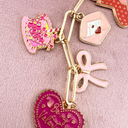 Valentine’s-themed charm keychain featuring pink and red enamel heart and love charms clipped to a phone case and displayed on a soft pink background.