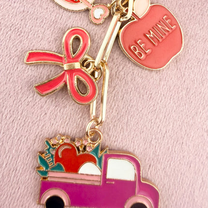 Valentine’s-themed charm keychain featuring pink and red enamel heart and love charms clipped to a phone case and displayed on a soft pink background.
