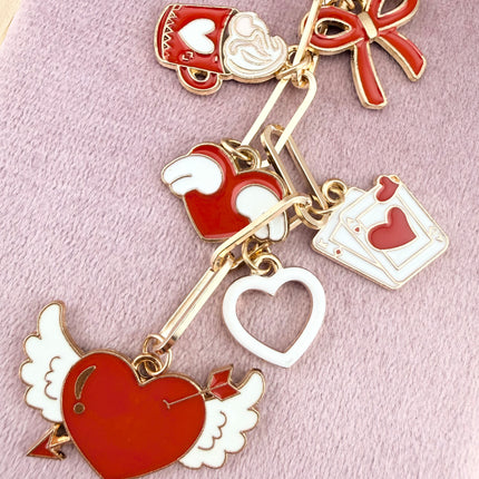 Valentine’s-themed charm keychain featuring pink and red enamel heart and love charms clipped to a phone case and displayed on a soft pink background.