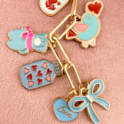 Valentine’s-themed charm keychain featuring pink and red enamel heart and love charms clipped to a phone case and displayed on a soft pink background.