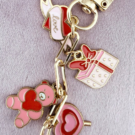 Valentine’s-themed charm keychain featuring pink and red enamel heart and love charms clipped to a phone case and displayed on a soft pink background.