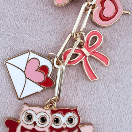 Valentine’s-themed charm keychain featuring pink and red enamel heart and love charms clipped to a phone case and displayed on a soft pink background.