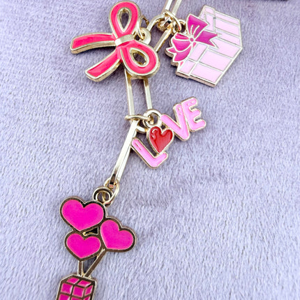 Valentine’s-themed charm keychain featuring pink and red enamel heart and love charms clipped to a phone case and displayed on a soft pink background.