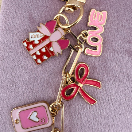 Valentine’s-themed charm keychain featuring pink and red enamel heart and love charms clipped to a phone case and displayed on a soft pink background.