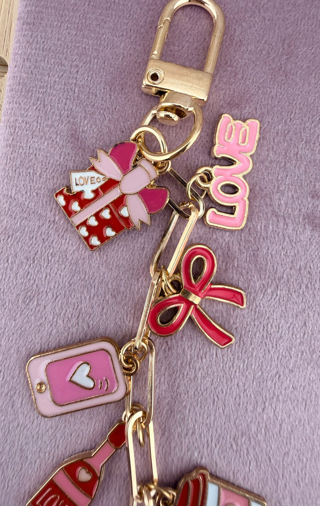 Valentine’s-themed charm keychain featuring pink and red enamel heart and love charms clipped to a phone case and displayed on a soft pink background.