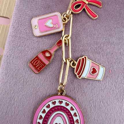Valentine’s-themed charm keychain featuring pink and red enamel heart and love charms clipped to a phone case and displayed on a soft pink background.