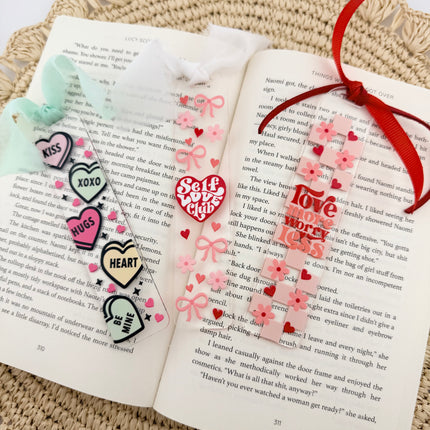 Clear acrylic Valentine’s Day bookmarks with pastel designs and soft chiffon ribbons resting inside open books, styled with a cozy, romantic bookish aesthetic.