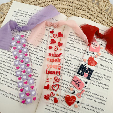 Clear acrylic Valentine’s Day bookmarks with pastel designs and soft chiffon ribbons resting inside open books, styled with a cozy, romantic bookish aesthetic.