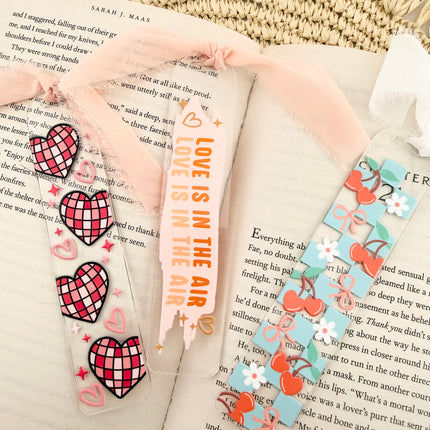 Clear acrylic Valentine’s Day bookmarks with pastel designs and soft chiffon ribbons resting inside open books, styled with a cozy, romantic bookish aesthetic.