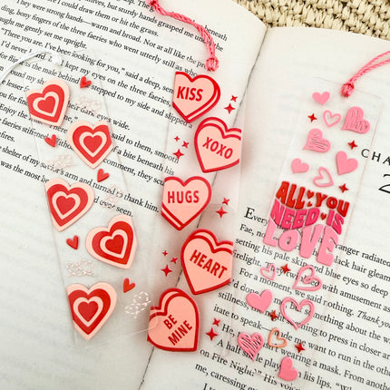 Handmade acrylic Valentine’s Day bookmarks featuring pink and red heart-themed designs with tassels, styled on open books for a cozy, festive reading aesthetic.