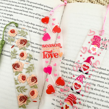 Handmade acrylic Valentine’s Day bookmarks featuring pink and red heart-themed designs with tassels, styled on open books for a cozy, festive reading aesthetic.