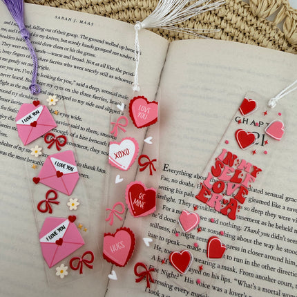 Handmade acrylic Valentine’s Day bookmarks featuring pink and red heart-themed designs with tassels, styled on open books for a cozy, festive reading aesthetic.