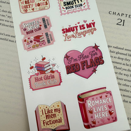 Multiple book lover sticker sheet designs featuring cozy reading themes, spicy book humor, hearts, books, and playful illustrations displayed on an open book background.