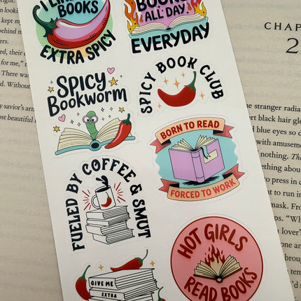 Multiple book lover sticker sheet designs featuring cozy reading themes, spicy book humor, hearts, books, and playful illustrations displayed on an open book background.