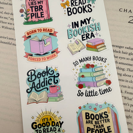 Multiple book lover sticker sheet designs featuring cozy reading themes, spicy book humor, hearts, books, and playful illustrations displayed on an open book background.
