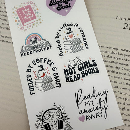Multiple book lover sticker sheet designs featuring cozy reading themes, spicy book humor, hearts, books, and playful illustrations displayed on an open book background.