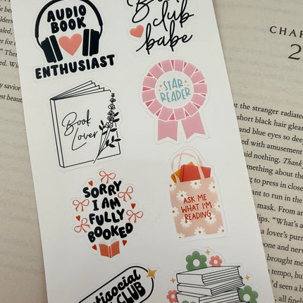 Multiple book lover sticker sheet designs featuring cozy reading themes, spicy book humor, hearts, books, and playful illustrations displayed on an open book background.