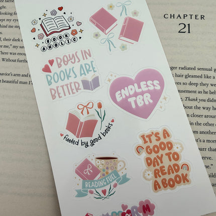 Multiple book lover sticker sheet designs featuring cozy reading themes, spicy book humor, hearts, books, and playful illustrations displayed on an open book background.