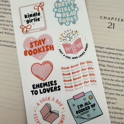 Multiple book lover sticker sheet designs featuring cozy reading themes, spicy book humor, hearts, books, and playful illustrations displayed on an open book background.