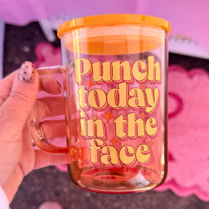 Clear mug with orange lid and text 'Punch today in the face' held by a hand against a pink background.