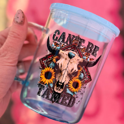 Clear mug with bull skull design and 'Can't Be Tamed' text, held by a hand against a pink background.