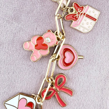 Valentine’s-themed charm keychain featuring pink and red enamel heart and love charms clipped to a phone case and displayed on a soft pink background.