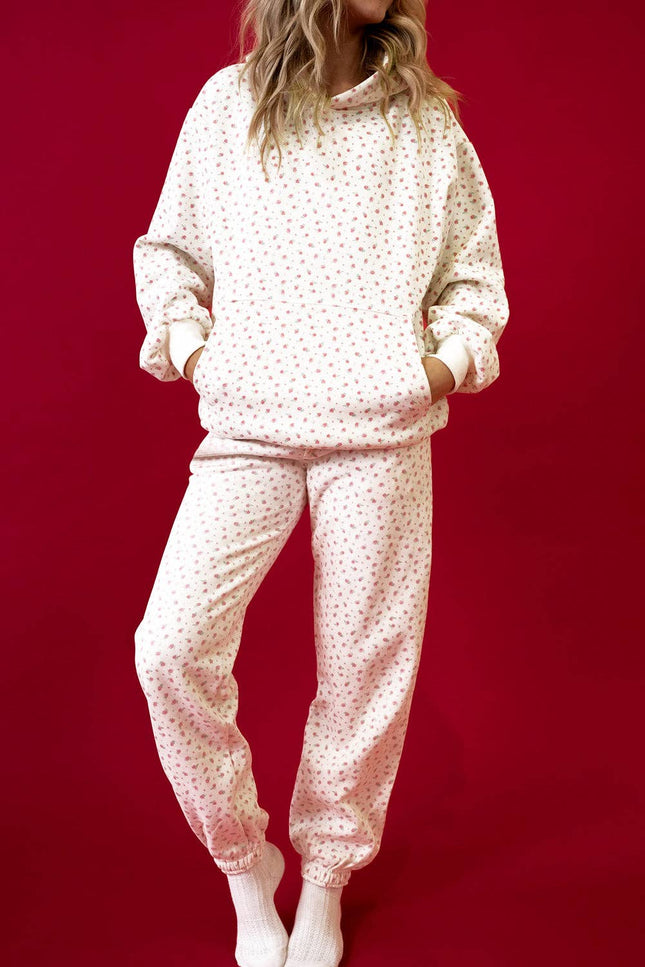 Model wearing a pink floral hoodie with a kangaroo pocket and matching sweatpants styled in a relaxed fit against a red background.